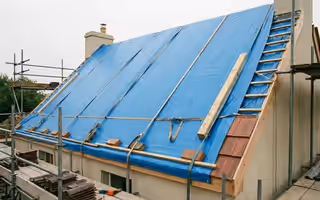 Toiture entièrement bâchée de bleu et sanglée, échafaudage monté, tuiles déposées empilées